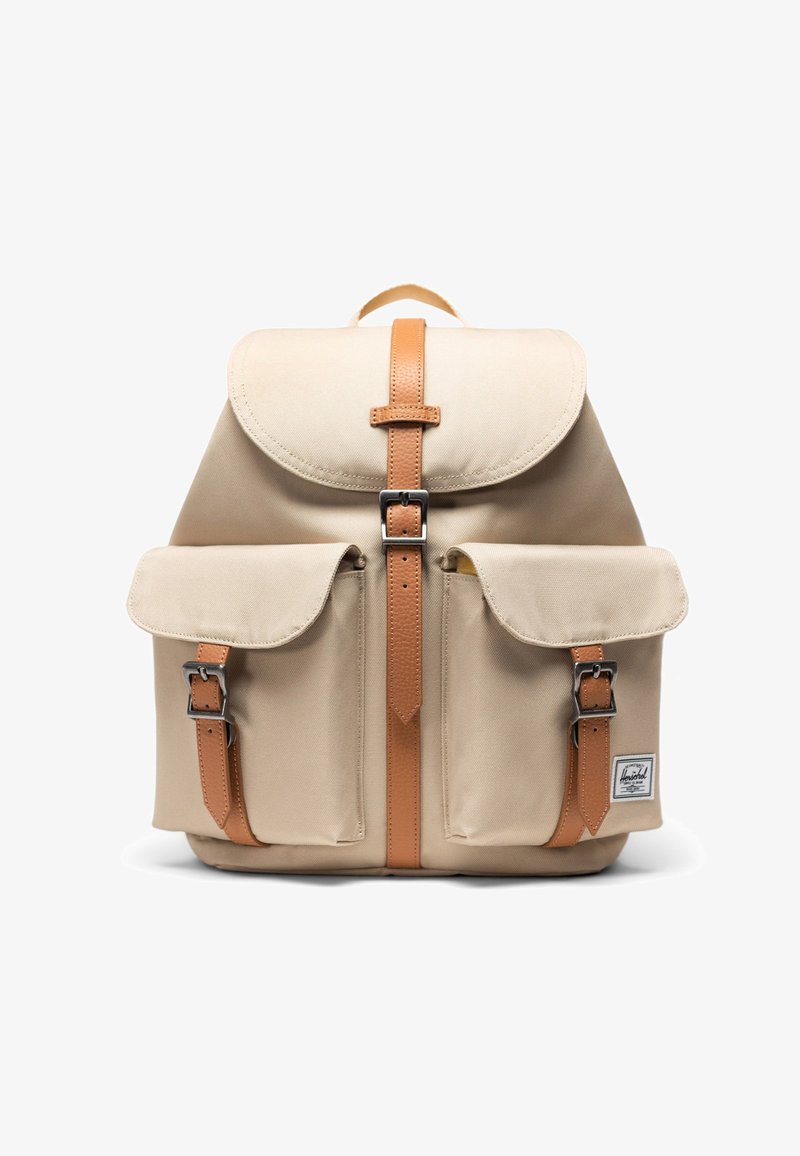 Sac à dos en toile beige avec des sangles et boucles en cuir fauve, deux poches avant, une poignée supérieure et une petite étiquette de marque sur la poche inférieure droite.