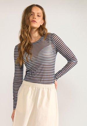 Jonge vrouw met lang golvend haar, gekleed in een bruin en blauw gestreept top met lange mouwen en een crèmekleurige rok, staand met één hand op haar heup.