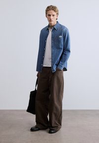 Μπλε πουκάμισο από τζιν με κουμπιά, φορεμένο πάνω από λευκό μπλουζάκι, σετ με καφέ παντελόνια με φαρδιά μπατζάκια. Μαύρη τσάντα tote και μαύρα παπούτσια ολοκληρώνουν την εμφάνιση.