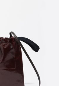 Bordowa skórzana torba ze sznurkiem o teksturowanym wykończeniu, z czarną materiałową rączką i smukłym brązowym paskiem.