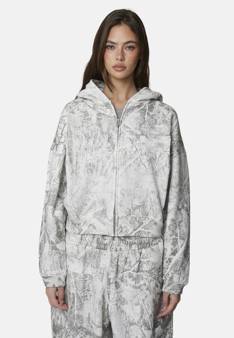 Kobieta w białej i szarej bluzie na zamek z abstrakcyjnym wzorem oraz dopasowanych spodniach, stojąca na tle jednolitego tła, patrząca przed siebie.