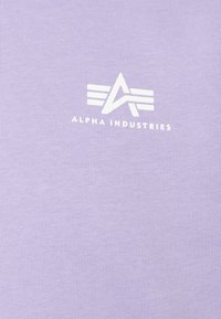Alpha Industries logo ar stilizētu "A" un trim horizontālām līnijām virs baltā teksta uz lilac auduma fona.
