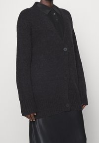 Sort strikket cardigan med V-hals, lange ærmer og ribbet kant. Har store sorte knapper og en blød, tekstureret stof.