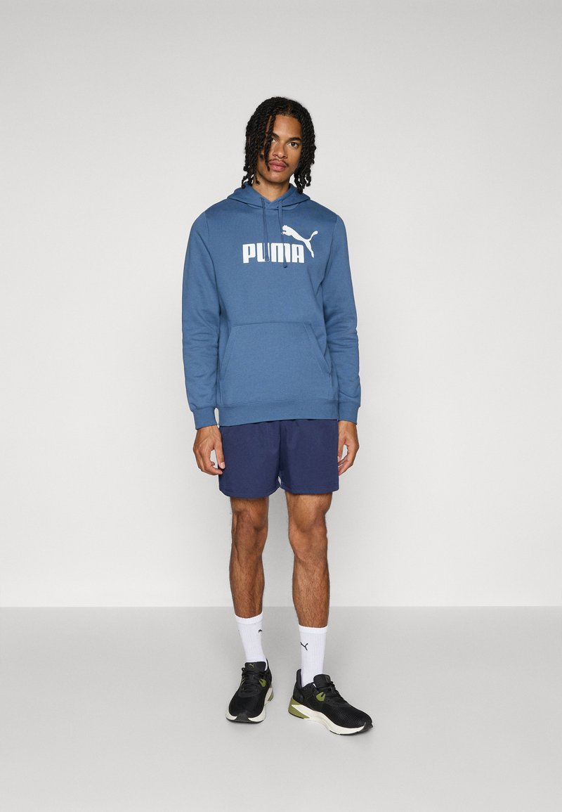 Blauwe hoodie met wit PUMA-logo, gecombineerd met marineblauwe shorts, witte sokken en zwarte sportschoenen, tegen een neutrale achtergrond.