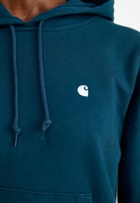 Teal cotton hoodie with a kangaroo pocket, braided drawstrings, and a small white logo on the left chest area. Soft texture and relaxed fit.