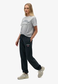 Camiseta de algodón gris con estampado "Superdry Athletic Essentials", combinada con pantalones de chándal negros con detalle del logo y cintura elástica. Zapatillas beige.