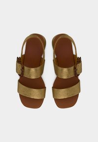 Ζευγάρι σανδάλια με χρυσό μεταλλικό λουράκι και καφέ επίπεδες σόλες, με αγκράφες και ανοιχτά δάχτυλα, σε λευκό φόντο.