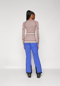 Langsleeve, getailleerde top van gemêleerd beige stof, gecombineerd met losse, felblauwe skibroeken. Opvallende donkere accenten bij de broekboorden.
