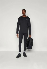 Svart langermet skjorte med logo, tettsittende svarte leggings og svarte joggesko. Modellen holder en svart ryggsekk, og viser et tilpasset atletisk utseende.