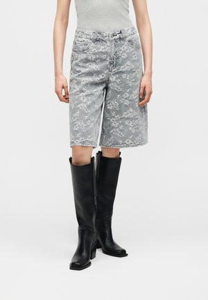 Personne portant un short en denim gris à fleurs, taille haute, associé à de grandes bottes noires en cuir à talons et un haut sans manches gris clair.