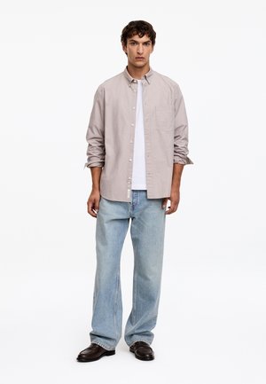 Homme portant une chemise beige clair à boutons sur un t-shirt blanc, un jean large bleu clair et des chaussures marron foncé, debout devant un fond blanc.