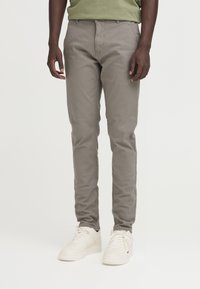 Pantaloni slim-fit grigi realizzati in tessuto misto cotone, con zip frontalmente, cinque tasche e cuciture a vista. Indossati con sneaker bianche.