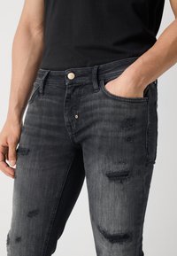 Jeans de mezclilla negra descolorida con detalles desgastados, cinco bolsillos, cierre frontal con botón y tela texturizada.