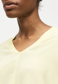 Εγγύς πλάνο του λαιμού και του κάτω μέρους του προσώπου ενός ατόμου που φοράει ένα ανοιχτό κίτρινο μπλουζάκι με V-λαιμόκοψη σε απλό λευκό φόντο.