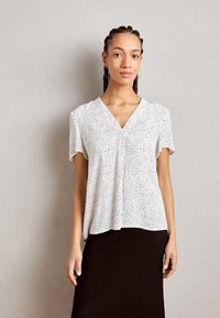 Blusa bianca a maniche corte con pois neri, design a V, tessuto morbido, silhouette leggermente drappeggiata, abbinata a una gonna scura.