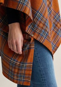 Rutet uldponcho i orange, blå og creme. Har knapdetaljer og en flowy drape, parret med mørk denim jeans.