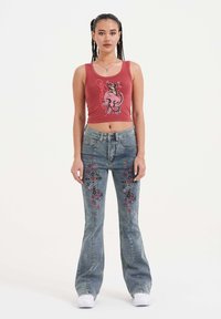 Top corto rojo con diseño gráfico y jeans acampanados azules que presentan bordados de flores y corazones. El modelo posa sobre un fondo claro.