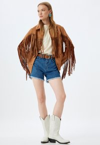 Franje bruine suède jas, crèmekleurig topje, denim shorts, bruine riem, witte cowboylaarzen met decoratieve stiksel, turkooize oorbellen.