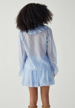 Kõrgetel hallil taustal seisev naine, kellel on lokkis juuksed, seljas läbipaistev helesinine pluus ja sobiv seelik, pildil näoga eemale vaadates.