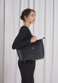 Schwarze Tragetasche mit glatter Textur, silbernen Beschlägen und einem Logo auf der Vorderseite, getragen von einem Modell vor einem neutralen Hintergrund.