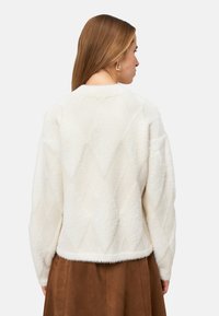 Pull en fausse fourrure crème avec motif en losange. Présente un col rond et des manches froncées. L'ourlet se situe au-dessus de la hanche.