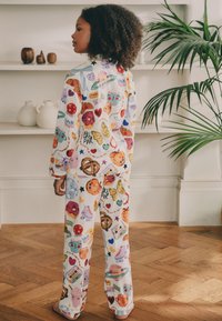 Ensemble de pyjama blanc avec un motif coloré et ludique comprenant des beignets, des cœurs, des papillons et d'autres illustrations funky. Tissu doux, coupe décontractée.