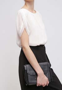 Blusa bianca e trasparente a maniche corte abbinata a pantaloni neri a vita alta. Il modello tiene una pochette in pelle nera texture con chiusura a pattina.