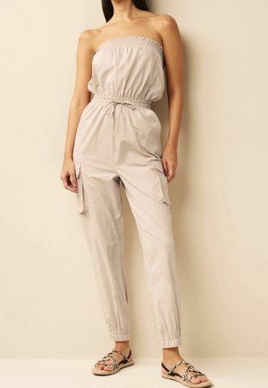 Femme portant une combinaison beige sans bretelles avec poches avant et poignets élastiques, associée à des sandales noires et blanches à brides tressées.