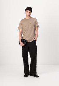 Beige t-skjorte med kort erme og lomme, med gult logo, kombinert med løse sorte bukser og svarte sko, en svart clutch holdt i hånden.