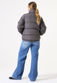 Doudoune à motif chevrons sombre, avec un col haut et des épaules arrondies, associée à un jean bleu ample et des chaussures noires.