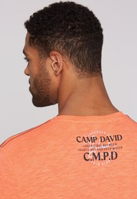 Camiseta de manga corta naranja con cuello redondo, que presenta un estampado de logo negro y costuras en negro contrastantes en los hombros.