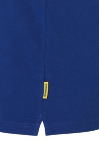 Blauwe gestructureerde stof, rechthoekige vorm, met aan de zijkant een klein geel label met #IAMNOMAD, en een splitzoom aan de onderkant.