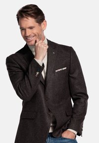 Donkerbruin wollen blazer met een getailleerde pasvorm, voorzien van een opgestikte zak met bloemenstof en kleurrijke knopen op de mouwen. Draagt over een lichte trui.