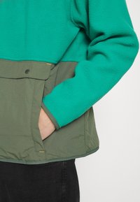 Zielona kurtka polarowa z kontrastowym oliwkowym brzegiem, posiadająca boczną kieszeń zapinaną na zatrzask oraz teksturowany, miękki materiał.