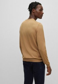 Maglione marrone lavorato a maglia con maniche lunghe e polsini a coste, abbinato a pantaloni blu navy. Texture liscia e scollatura a girocollo sono visibili.