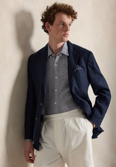 Άνδρας με κατσαρά κόκκινα μαλλιά, φορούντας μπλε σκούρα σακάκι, μπλε πουκάμισο με ρίγες και κουμπιά, άσπρο παντελόνι και μαντήλι με πουά στην τσέπη, στέκεται μπροστά σε μπεζ τοίχο.