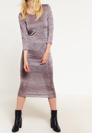 Vrouw draagt een mauve jurk met lange mouwen en een lengte tot de knieën, gecombineerd met zwarte enkelboots, staande tegen een effen lichte achtergrond.