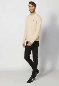 Beige geribde lange mouw trui met ronde hals, gedragen over zwarte slim-fit jeans en zwarte sneakers, met een subtiele logodetail op de borst.