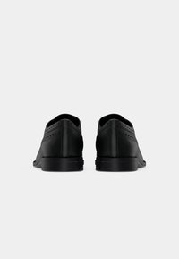 Paire de chaussures habillées en cuir noir avec des détails brogue montrées de dos sur un fond clair uni.