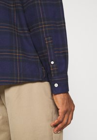 Chemise en flanelle à carreaux navy avec des lignes orange et verte, dotée d'un poignet à boutons et d'un tissu texturé, associée à un pantalon kaki.