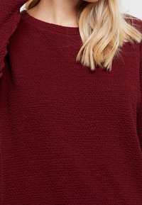 Burgundské texturované svetr s kulatým výstřihem a dlouhými rukávy, s jemným, mírně vyvýšeným vzorem na povrchu.