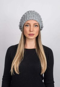 Šedá pletená čepice s texturovaným, žebrovaným designem, nošená přes rovné vlasy. Na neutrálním pozadí. Černý top viditelný dole.