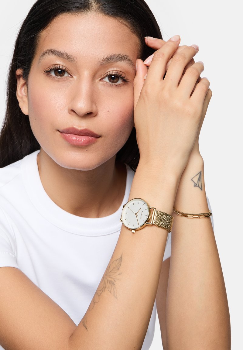 Goldene Uhr mit weißem Ziffernblatt und strukturiertem Mesh-Armband, getragen an einem Handgelenk neben einem schlanken goldenen Armband. Der Hautton ist gebräunt und der Arm hat ein Tattoo.