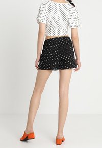 Femme portant un crop top blanc à pois noirs, un short à pois noirs, et des chaussures à talons carrés orange, se tenant de dos.