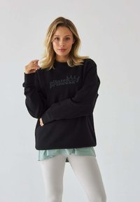 Czarny sweter z logo "Pilates Princess" w kolorze szarym. Posiada okrągły dekolt i luźny krój, w połączeniu z jasnoniebieską bluzką i białymi legginsami.