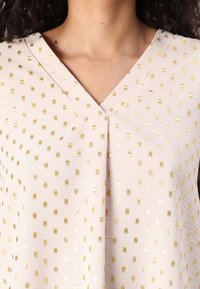 Blusa rosa chiaro con scollo a V, caratterizzata da un tessuto testurizzato decorato con piccoli pattern quadrati dorati sparsi.