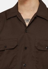 Osoba oblečená v tmavě hnědé košili na knoflíky se dvěma kapsami na klopě a s viditelným tetováním oka na horním hrudníku pod límcem.