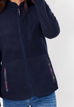 Veste en polaire bleu marine avec des tirettes de fermeture éclair à motif floral sur la poitrine et les poches latérales, portée avec un jean bleu.