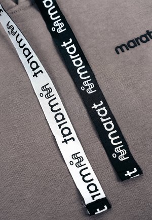 Twee grijze capuchonkoorden, één zwart en één wit, elk bedrukt met het woord "tamart" en een klein gestileerd figuurtje, op een grijze stoffen achtergrond.