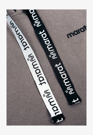 Twee grijze capuchonkoorden, één zwart en één wit, elk bedrukt met het woord "tamart" en een klein gestileerd figuurtje, op een grijze stoffen achtergrond.